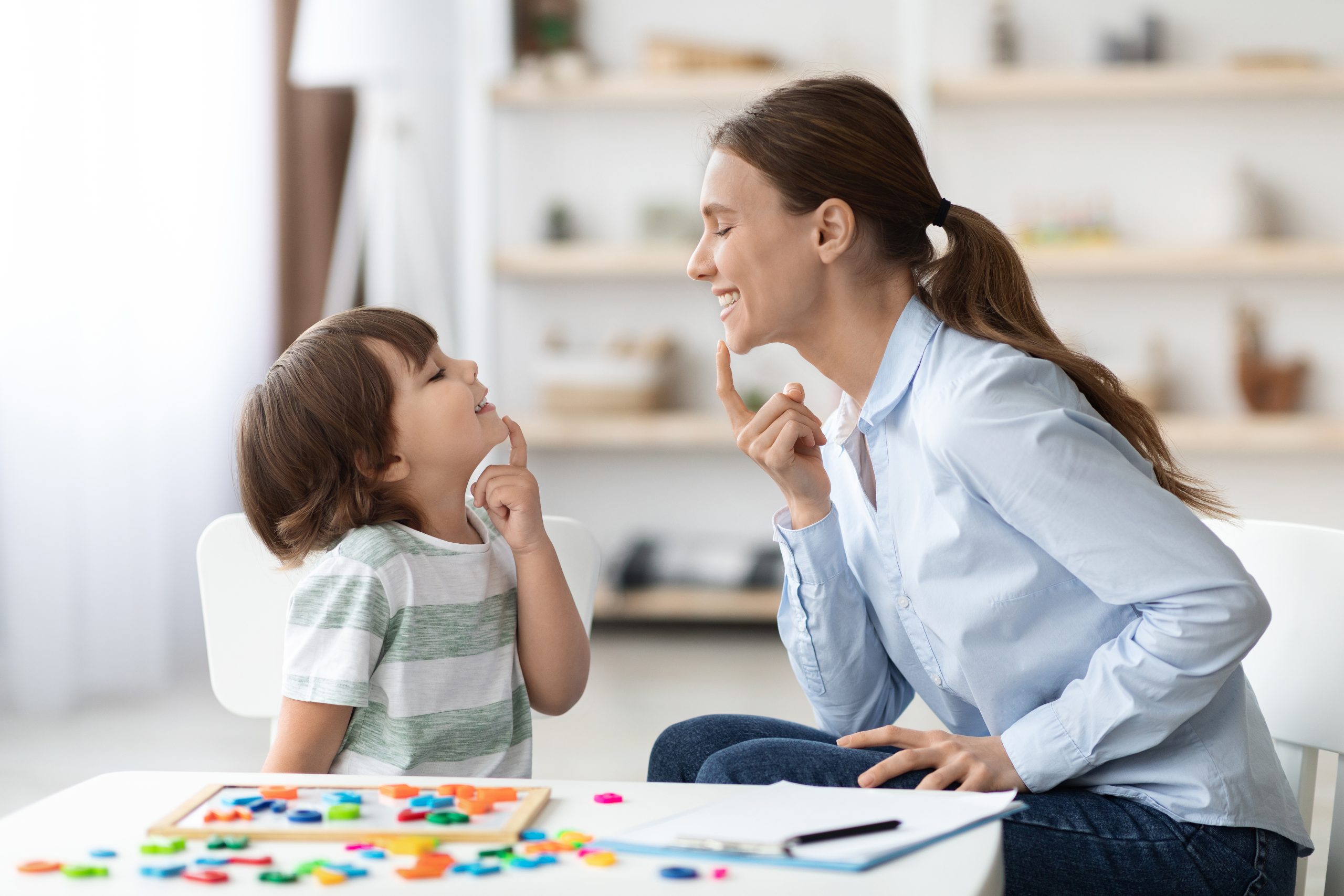 Speech training for kids. Professional woman training with little boy at cabinet, teaching right articulation exercises Speech training for kids. Professional woman specialist training with little boy at cabinet, teaching him right articulation exercises, side view
