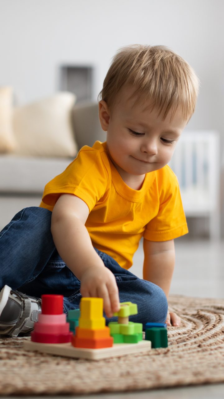 Adorable little boy playing with educational wooden toy at home, sitting on floor carpet in living room, copy space Adorable little boy playing with educational wooden toy at home, sitting on floor carpet in living room interior, copy space. Toddler with wooden colorful stacking and sorting toy. Child development